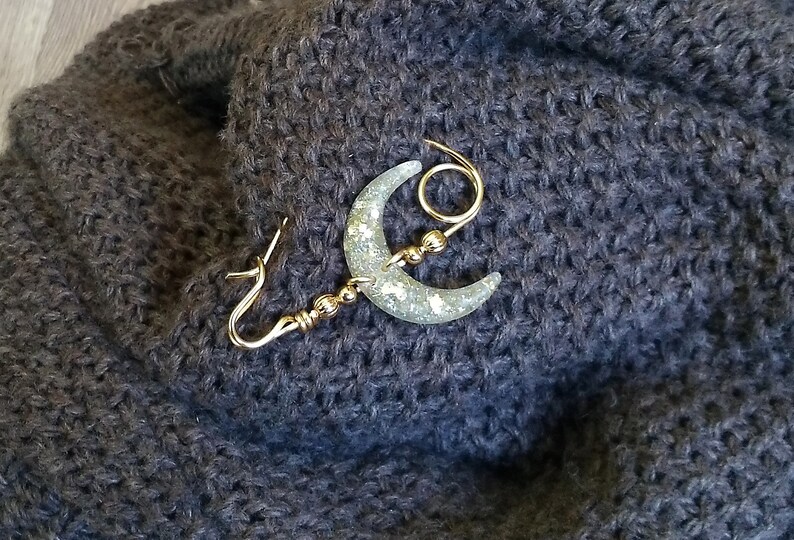 May include: A gold crescent moon brooch with glitter embedded in the resin. The brooch has a decorative loop and a pin closure.