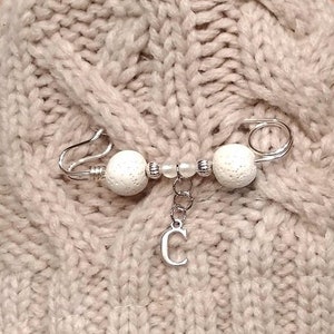 May include: A silver safety pin with white beads and a silver charm with the letter C hanging from it.