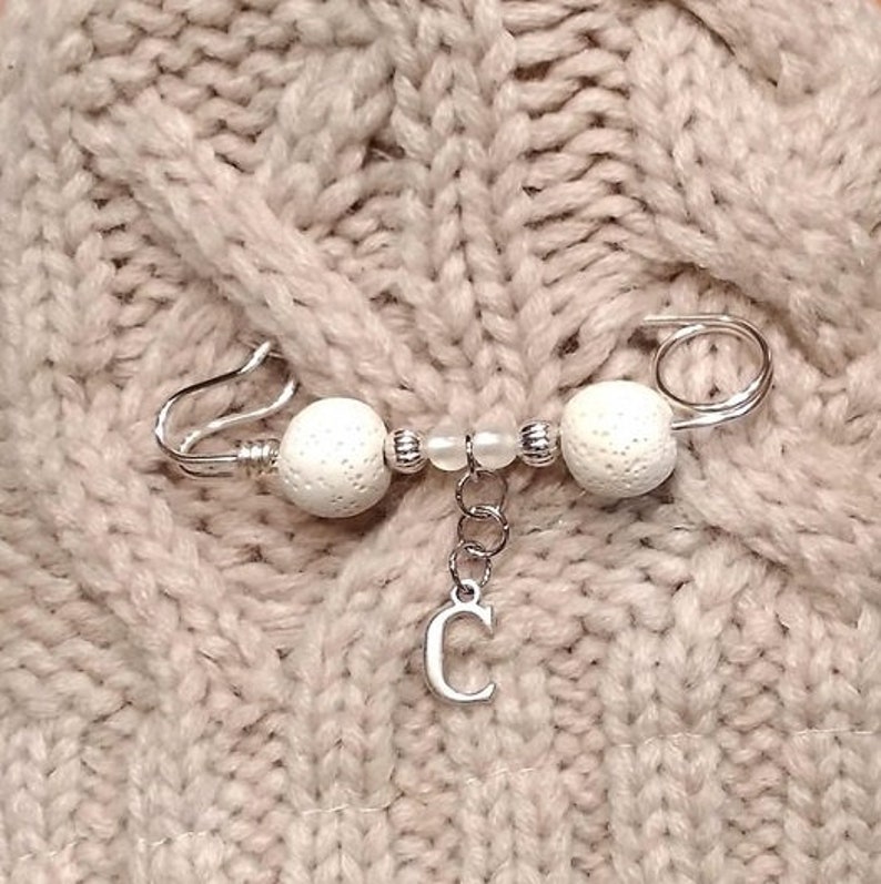 May include: A silver safety pin with white beads and a silver charm with the letter C hanging from it.