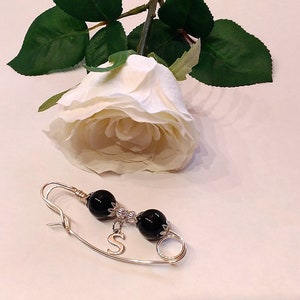 May include: A gold safety pin with black beads and a silver charm with the letter 'S'.