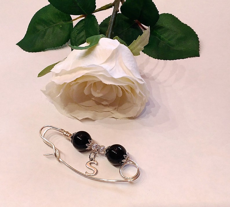 May include: A gold safety pin with black beads and a silver charm with the letter 'S'.