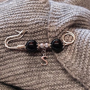 May include: A silver brooch with two black beads and a silver charm with the letter 'S' hanging from it. The brooch is pinned to a gray knitted fabric.