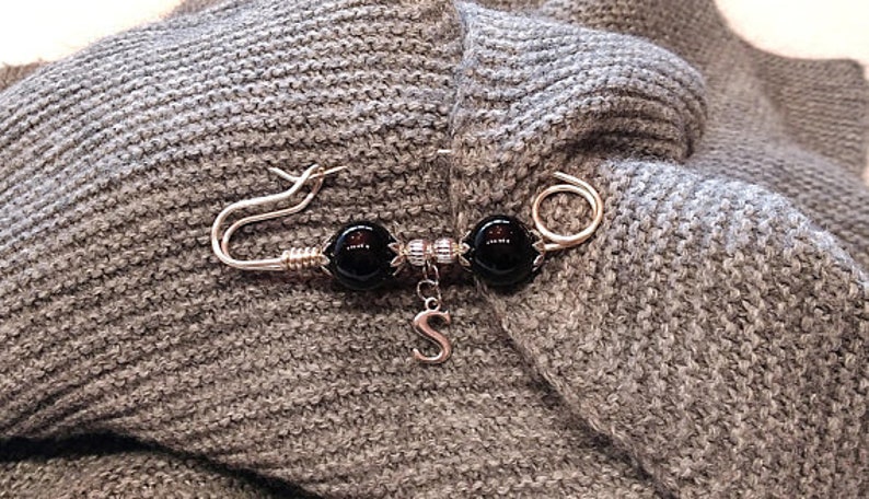 May include: A silver brooch with two black beads and a silver charm with the letter 'S' hanging from it. The brooch is pinned to a gray knitted fabric.