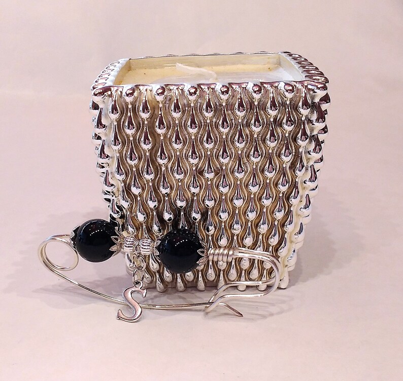 May include: A silver candle holder with a textured design resembling raindrops. The holder is filled with a white candle. A silver brooch with black beads is in the foreground.