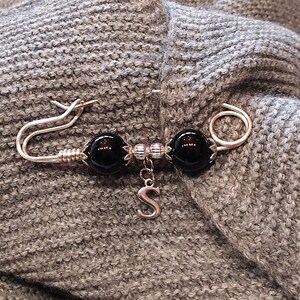 May include: A silver safety pin with two black beads and a silver charm with the letter 'S' hanging from it. The pin is attached to a gray knitted fabric.