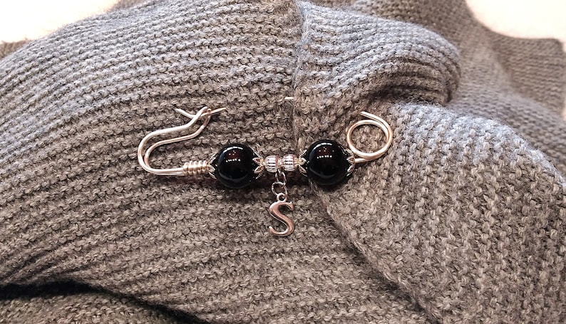 May include: A silver safety pin with two black beads and a silver charm with the letter 'S' hanging from it. The pin is attached to a gray knitted fabric.