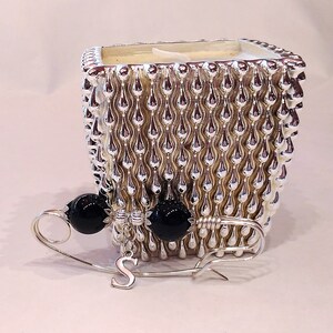 May include: A silver candle holder with a textured design and black beads attached to a safety pin with a letter 'S' charm.