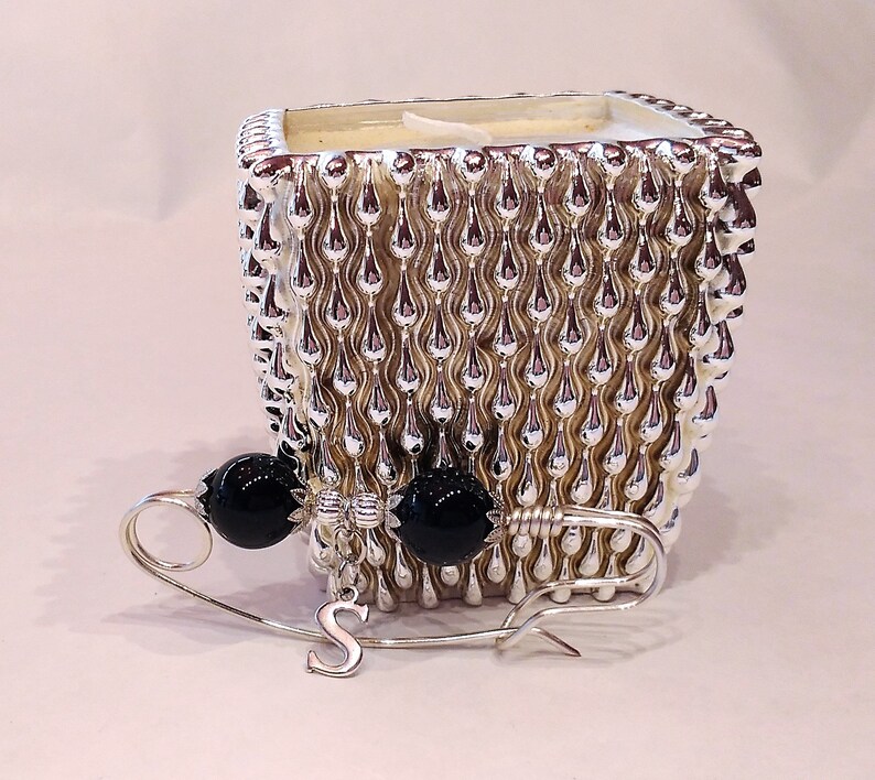 May include: A silver candle holder with a textured design and black beads attached to a safety pin with a letter 'S' charm.