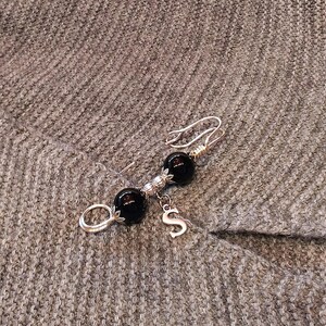May include: A silver brooch with two black beads and a silver charm with the letter 'S'.