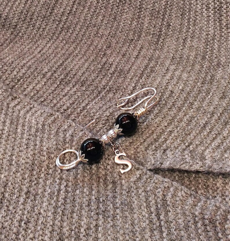 May include: A silver brooch with two black beads and a silver charm with the letter 'S'.