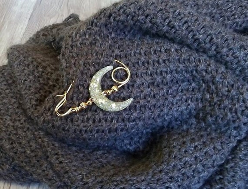 May include: A gold crescent moon brooch with glitter accents on a dark gray knitted fabric.