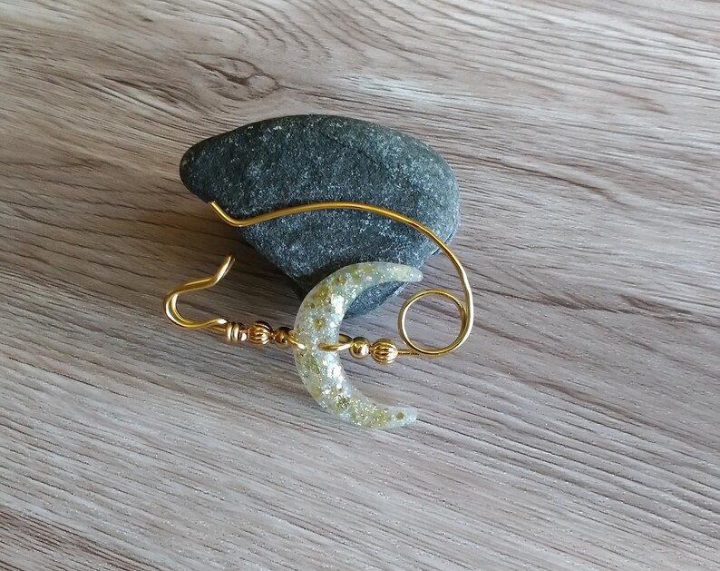 May include: A gold wire crescent moon with glitter accents, attached to a gold wire loop. The moon is hanging from a gray rock.