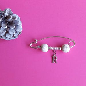 May include: A silver safety pin with white beads and a silver charm with the letter 'R' hanging from it. The pin is on a pink background.