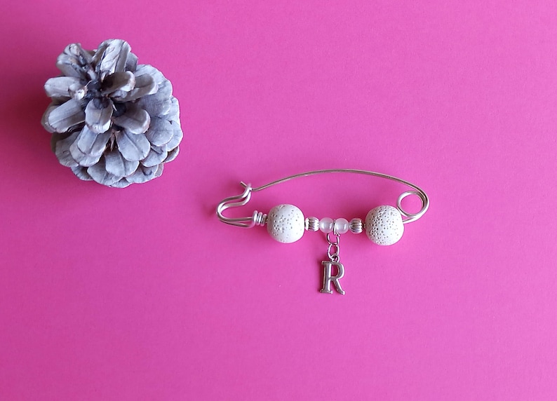 May include: A silver safety pin with white beads and a silver charm with the letter 'R' hanging from it. The pin is on a pink background.