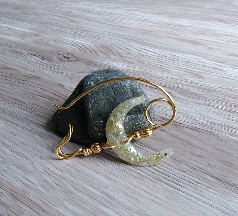 May include: A gold wire crescent moon charm with a white glitter finish. The charm is attached to a gold wire loop and has a gold bead accent.