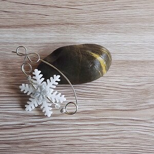 May include: A silver wire snowflake ornament with a white snowflake charm. The ornament is on a wooden surface with a dark grey stone.
