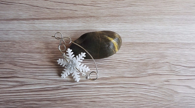May include: A silver wire snowflake ornament with a white snowflake charm. The ornament is on a wooden surface with a dark grey stone.