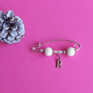 May include: A silver safety pin with white beads and a silver charm with the letter 'R' hanging from it.