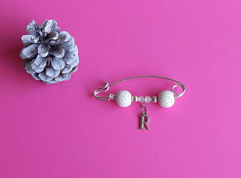 May include: A silver safety pin with white beads and a silver charm with the letter 'R' hanging from it.