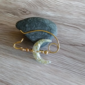 May include: A gold wire crescent moon with glitter accents, attached to a gold wire loop. The moon is hanging from a gray rock.