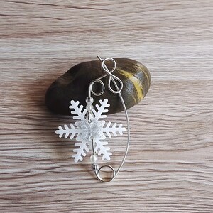 May include: A silver snowflake charm with white beads hanging from a silver wire loop. The charm is resting on a brown and yellow stone.