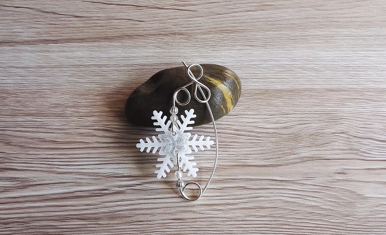 May include: A silver snowflake charm with white beads hanging from a silver wire loop. The charm is resting on a brown and yellow stone.