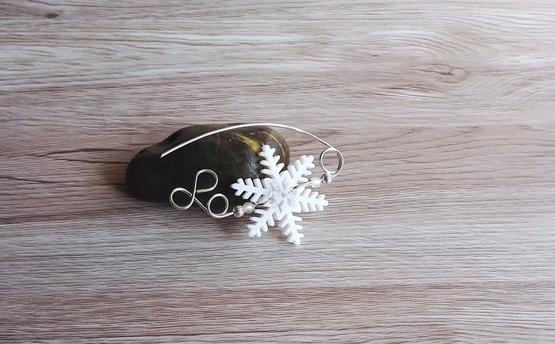 May include: A white snowflake charm attached to a silver wire with a silver loop. The charm is on a brown rock.