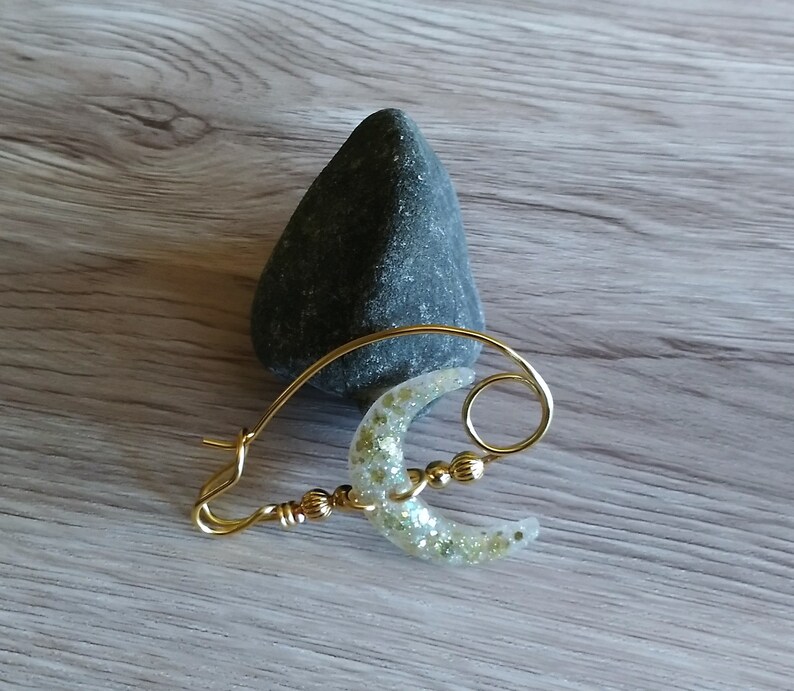 May include: Gold wire hoop earring with a crescent moon shaped charm. The charm is made of clear resin with gold glitter embedded in it.