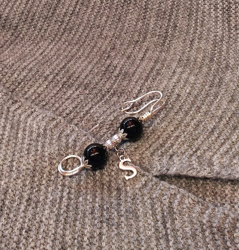 May include: A silver brooch with two black beads and a silver charm with the letter 'S' hanging from it.