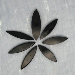 May include: A collection of seven dark, elongated, oval-shaped objects arranged in a circular pattern, resembling a flower. Each object has two small, silver-colored rivets. The objects are likely decorative or functional elements.