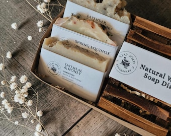 Peut inclure: Un coffret cadeau contenant trois savons étiquetés Lavender Honey, Spring Equinox et Oatmeal & Honey, ainsi qu'un porte-savon en bois. Chaque savon est enveloppé d'une bande de papier blanc avec le logo Stay Wild Apothecary imprimé.