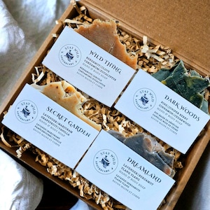 May include: A brown cardboard gift box filled with four bars of handmade soap. Each soap bar is labelled with its scent and ingredients, including "Wild Thing" and "Secret Garden." The soaps are surrounded by shredded paper.