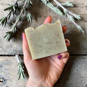 May include: A light green, rectangular bar of soap with visible flecks is held in a hand with purple nail polish. Sprigs of green herbs are arranged around the soap on a wooden surface. The soap appears to be handmade.