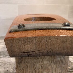 May include: A handcrafted wooden candle holder with a natural wood grain finish. The holder features two circular openings and a metal strip secured with screws. The wood has a warm, reddish-brown hue.