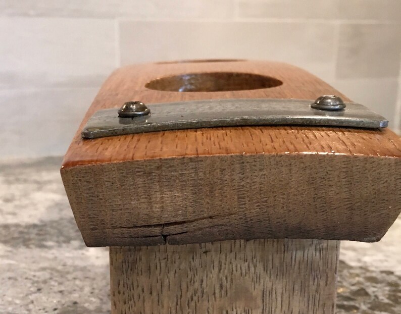 May include: A handcrafted wooden candle holder with a natural wood grain finish. The holder features two circular openings and a metal strip secured with screws. The wood has a warm, reddish-brown hue.