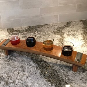 May include: A wooden beer flight with four small glasses, each filled with a different colored beer. The wooden tray has a curved design and metal accents. The beers range in color from light amber to dark brown.