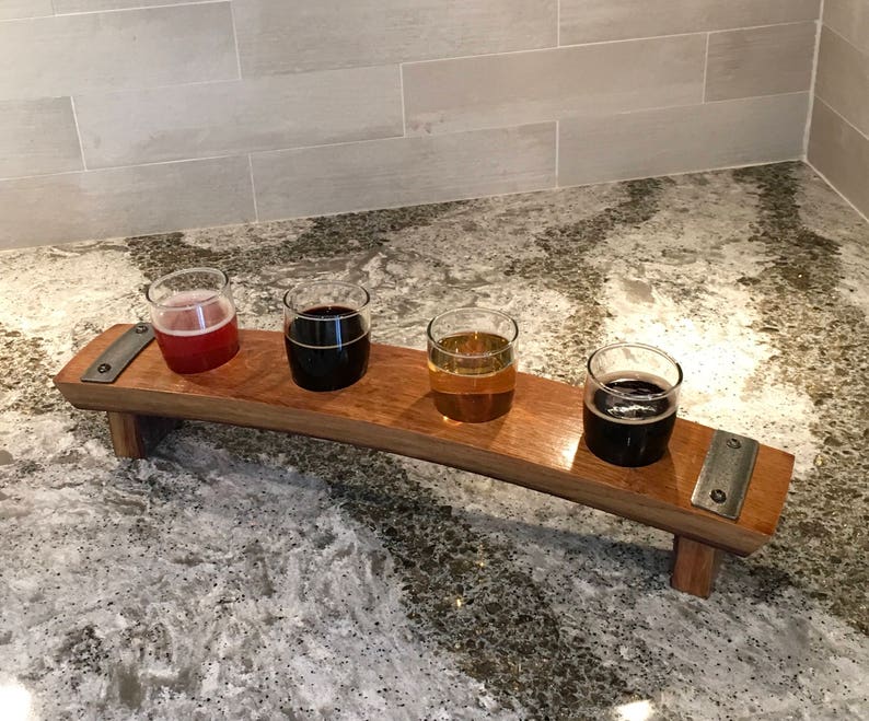 May include: A wooden beer flight with four small glasses, each filled with a different colored beer. The wooden tray has a curved design and metal accents. The beers range in color from light amber to dark brown.