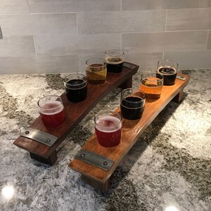 May include: Two wooden beer flight boards, each holding three glasses of beer. The beers vary in color, from light amber to dark brown. The boards have a curved design and are accented with metal brackets.