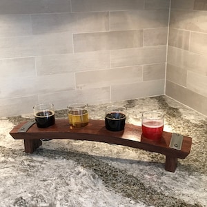 May include: A wooden beer flight with four glasses, each filled with a different colored beer. The wooden tray has a curved design and metal accents. The beers range in color from dark brown to light red.
