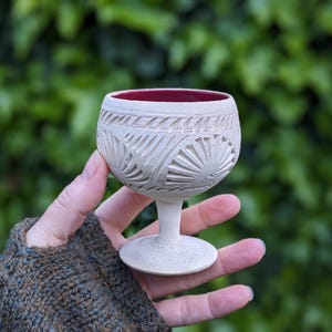 May include: A small, off-white ceramic wine glass with a carved design. The interior of the glass is a deep red color. The glass is held in a hand against a blurred green background.