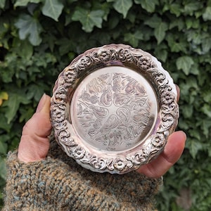May include: A small, round, silver-colored coaster with an ornate floral and scroll design. The coaster has a detailed, raised border and a central design of stylized leaves. The background is a blurred view of green foliage.