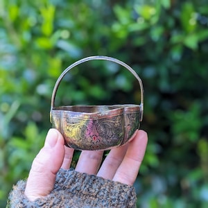 May include: A small, round, metallic basket featuring an intricate, dark, engraved floral pattern across its exterior surface. The vessel includes a thin, arched handle attached to the sides, with a scalloped rim and a polished, reflective finish.