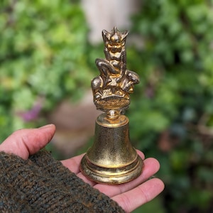 May include: A gold-coloured bell with a detailed figurine on top, held in a hand. The figurine depicts a seated figure with a crown. The bell has a flared base and a textured surface. The background is blurred green foliage.