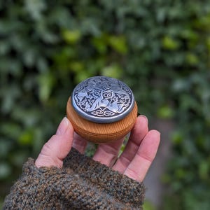 May include: A round wooden box with a silver-colored lid featuring an intricate Celtic design. The box is held in a hand, with a blurred green background of foliage. The lid has a detailed pattern of animals and knotwork.