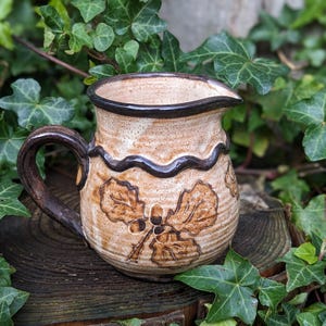 May include: A handmade ceramic pitcher with a brown handle and spout. The beige pitcher features a dark brown rim and a wavy design. Oak leaf and acorn designs are etched on the front. The pitcher is sitting on a wooden surface surrounded by green ivy.