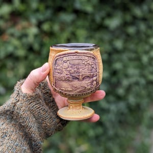 May include: A small, tan-colored ceramic goblet with a detailed relief of a landscape scene, possibly a bridge and buildings, labeled "Yorkshire." The goblet has a pedestal base and a dark rim.