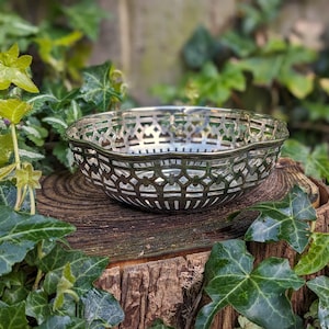 May include: A silver-toned, openwork bowl with a decorative geometric pattern. The bowl is placed on a weathered wooden surface, surrounded by green foliage. The bowl's intricate design is a prominent feature.