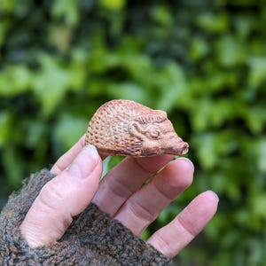 May include: A small, brown ceramic hedgehog figurine with detailed texture. The hedgehog is held in a hand against a blurred green background. The figurine has a detailed face and a textured body.
