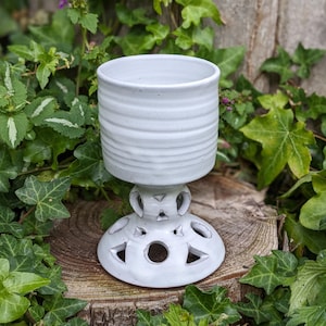 May include: A white ceramic pedestal planter with a cylindrical top and a decorative base featuring cut-out shapes. The planter is surrounded by green foliage and sits on a wooden surface. The top has horizontal ridges.