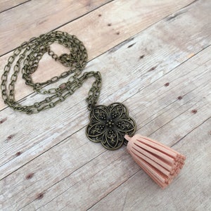 May include: A delicate necklace featuring a bronze chain and an intricate filigree pendant. The pendant is adorned with a blush pink tassel.